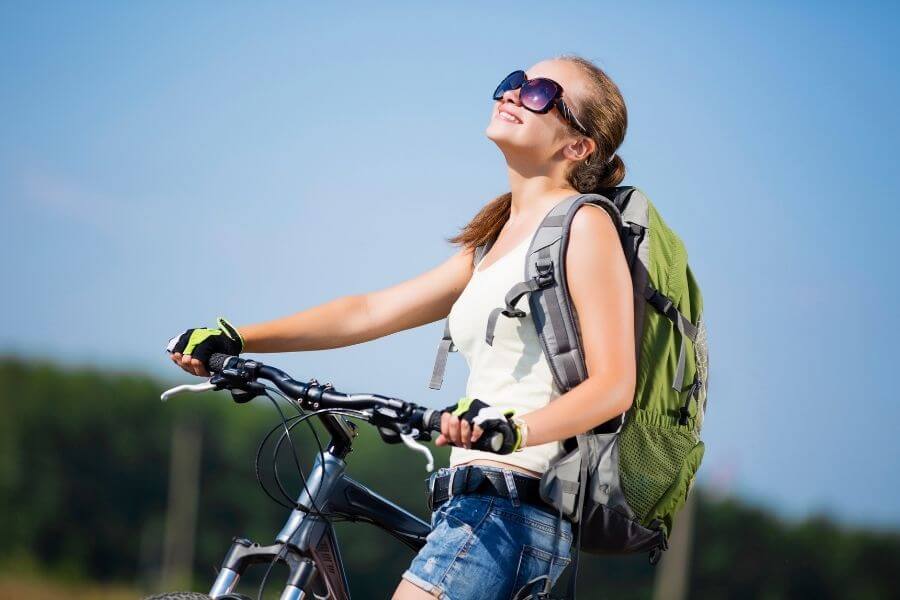 Smiling young girl with backpack and bicycle along roadside on a sunny day having positive mind